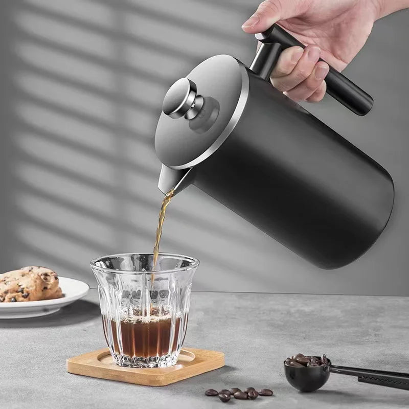 Person pouring coffee from a black French press into a glass on a gray surface with cookies and coffee beans.