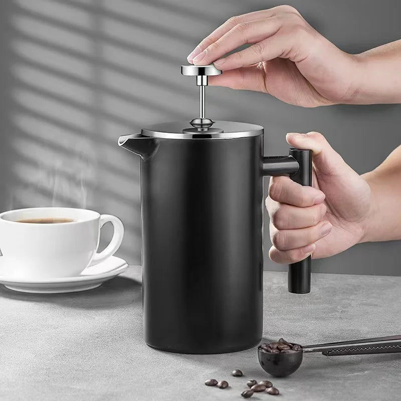 Person using a black French press to make coffee with a cup of coffee and coffee beans on a gray surface.