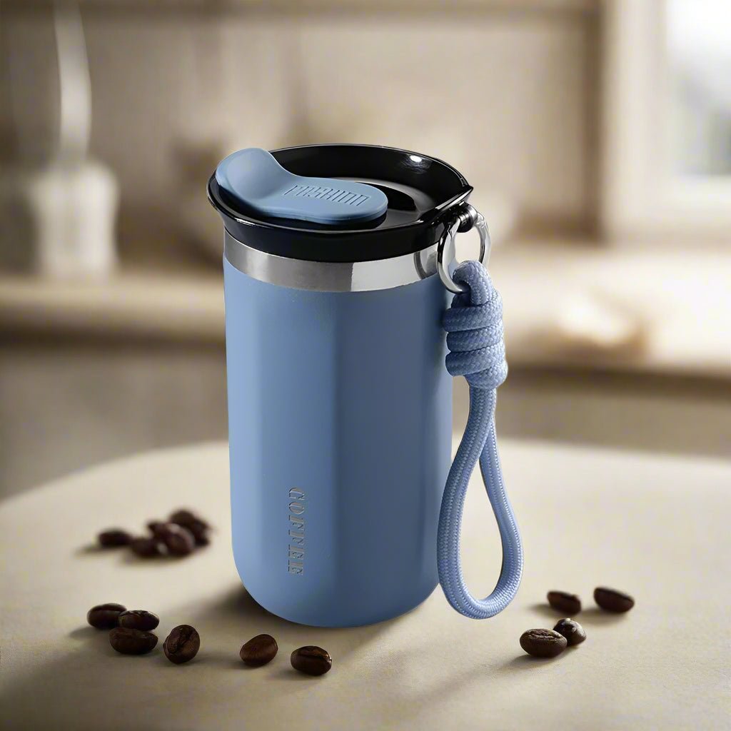 Blue tumbler with a black lid and rope handle on a surface with coffee beans.