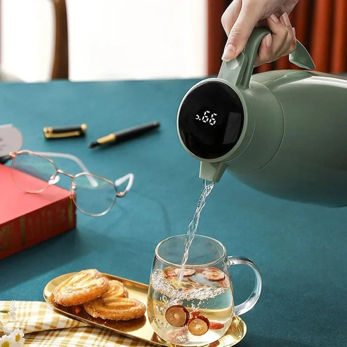Person pouring water from a digital Thermal Coffee Carafe into a glass mug on a table with a book and glasses.