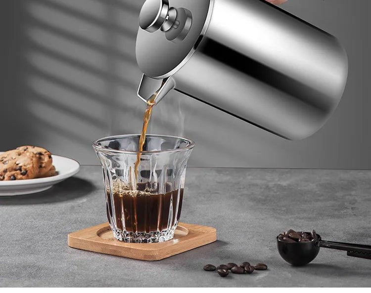 Stainless steel kettle pouring coffee into a glass on a gray surface with cookies and coffee beans.