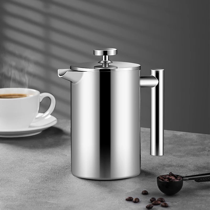 Stainless steel French press on a gray surface with a cup of coffee and coffee beans in the background.