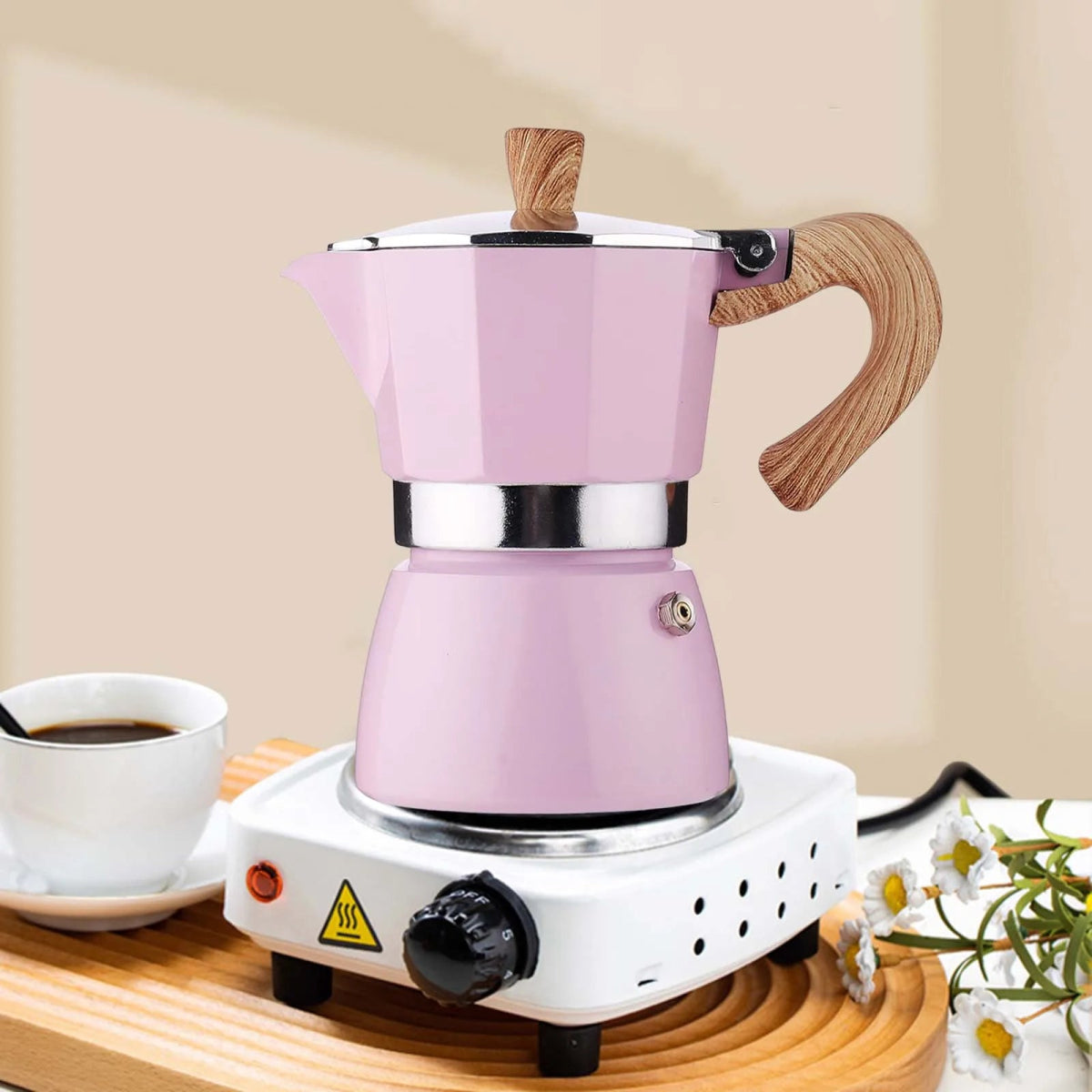 Pink espresso maker on a wooden stand with a cup of coffee and flowers in the background.