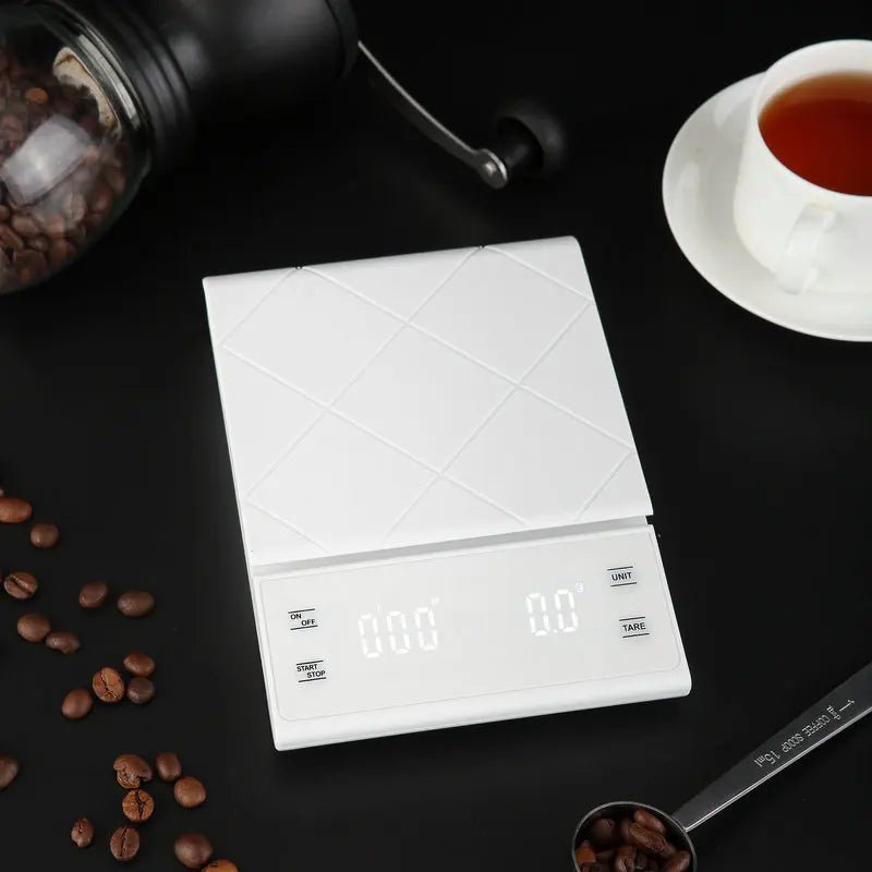 White digital kitchen scale on a dark surface with coffee beans and a cup of tea.