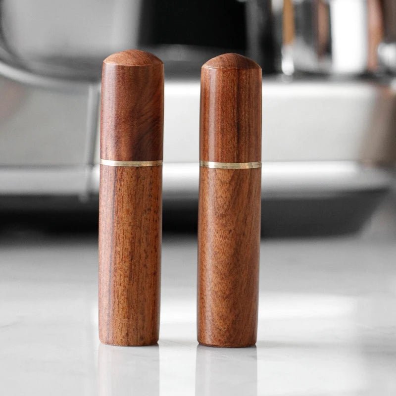 Two wooden pepper grinders with gold bands on a reflective surface.