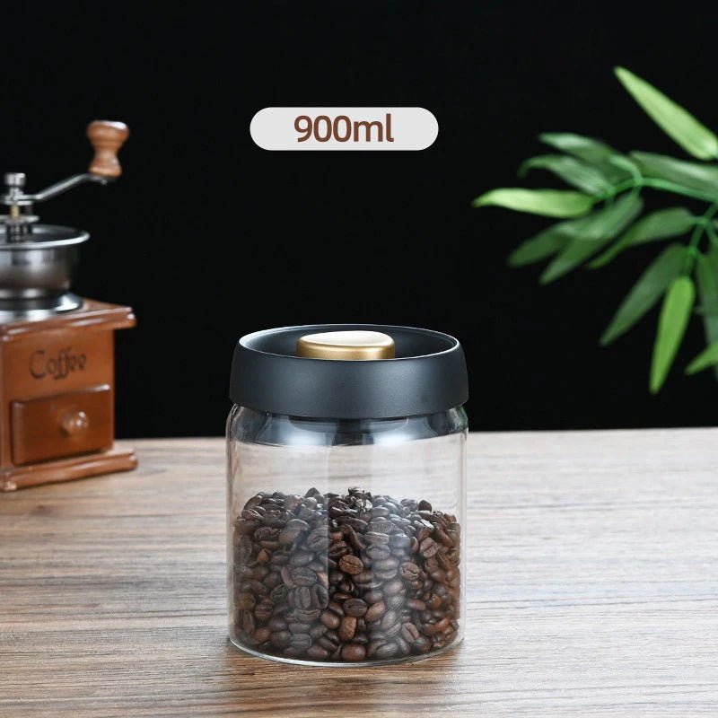 Glass jar with coffee beans and black lid on a wooden surface, with a coffee grinder and plant in the background.