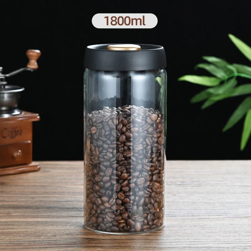Clear container with coffee beans and black lid on a wooden surface, with a coffee grinder and plant in the background.