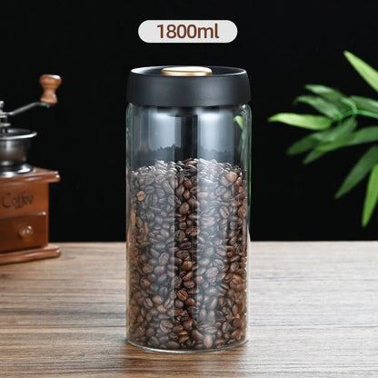 Clear container with coffee beans and black lid on a wooden surface, with a coffee grinder and plant in the background.