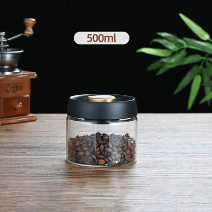 Glass container with coffee beans on a wooden surface, with a coffee grinder and plant in the background.