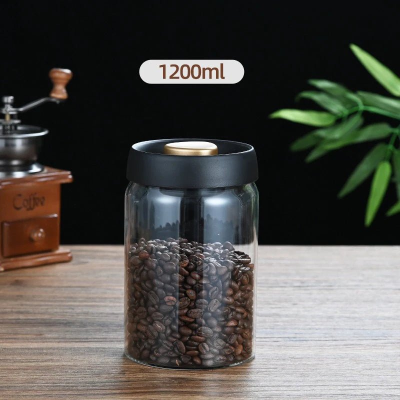 Glass jar with coffee beans and black lid on a wooden surface, with a coffee grinder and plant in the background.