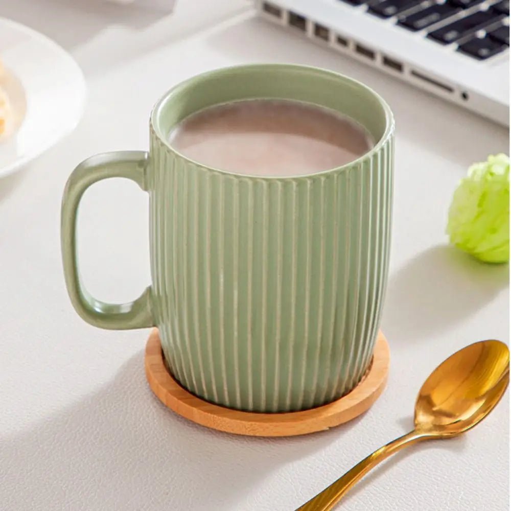 Green mug with striped pattern on a wooden coaster, filled with a hot beverage, on a light surface with a laptop and decorative items.
