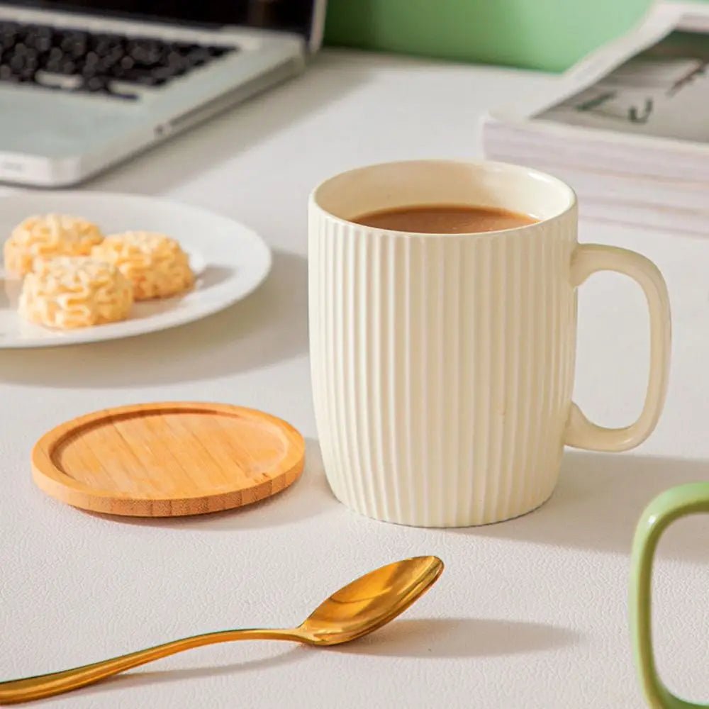 White ceramic mug with coffee, a wooden coaster, gold spoon, and cookies on a light surface.