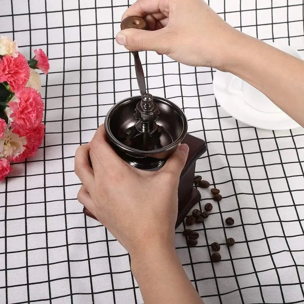 Person using a manual coffee grinder with coffee beans on a checkered tablecloth.