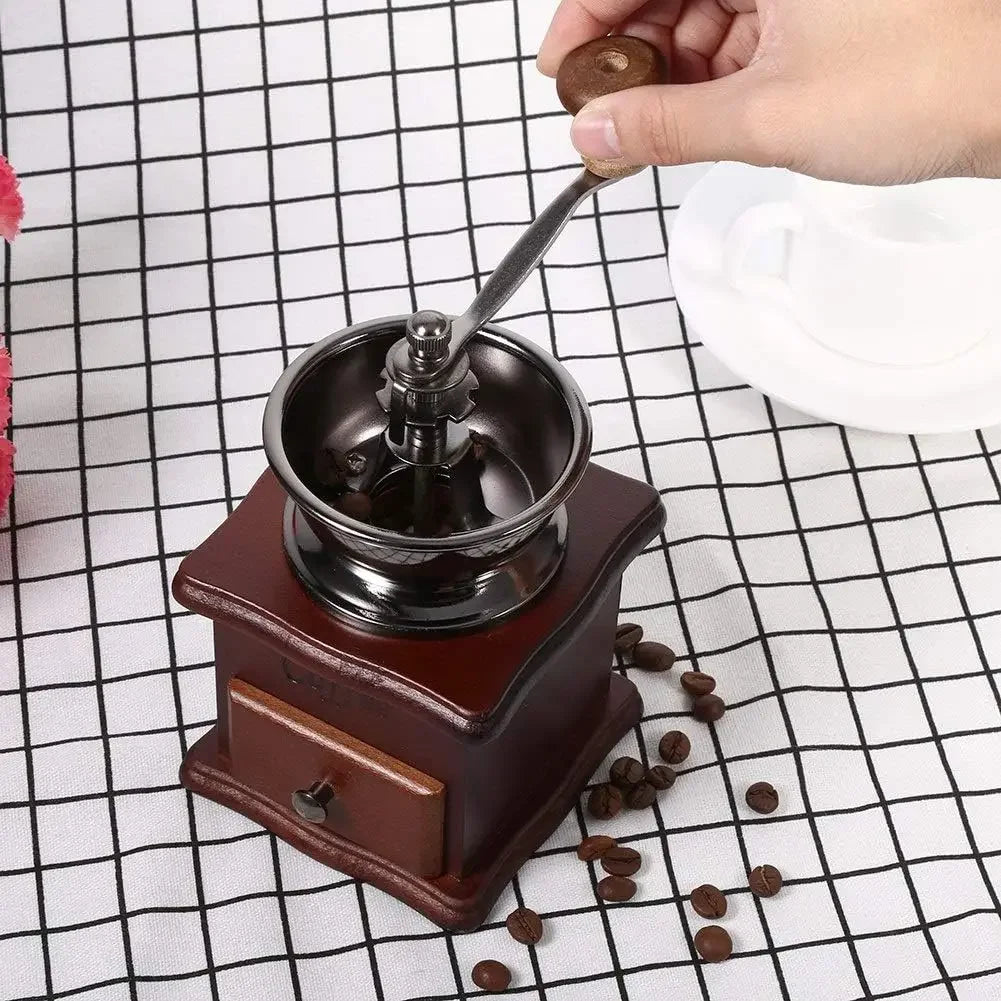 Hand using a manual coffee grinder with scattered coffee beans on a checkered tablecloth.