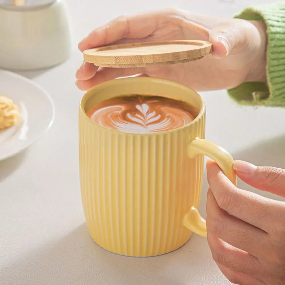 Yellow mug with a wooden lid being held by a person on a white surface.